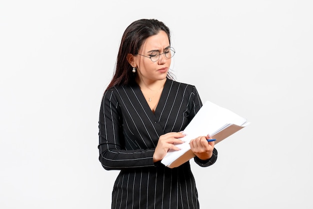 Vista frontal mujer joven en traje oscuro estricto sosteniendo y comprobando archivos sobre fondo blanco trabajo de oficina de negocios de documento femenino
