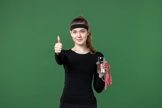 Vista frontal mujer joven con vaso de agua y medida de cintura sobre fondo verde deporte atleta mujer color de cuerpo salud entrenamiento negro