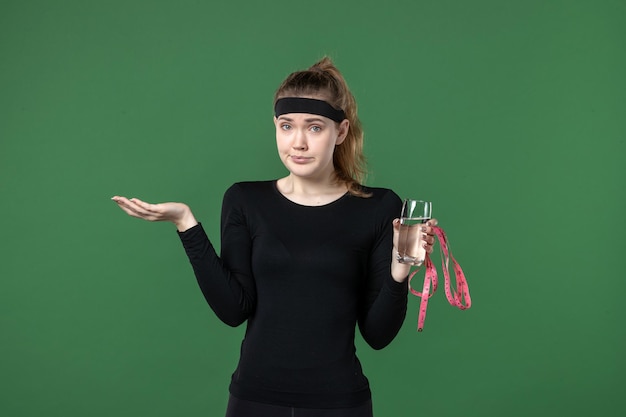 Vista frontal de la mujer joven con un vaso de agua y medir la cintura sobre fondo verde, cuerpo deportivo, salud, entrenamiento negro, color, mujer, atleta