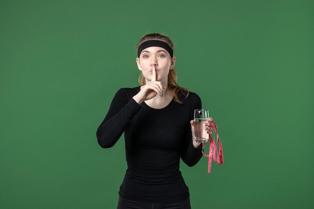 Vista frontal de la mujer joven con un vaso de agua y medir la cintura sobre fondo verde, salud, deporte, atleta, mujer, cuerpo, color, negro, entrenamiento