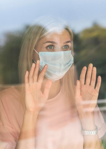 Vista frontal de la mujer con máscara médica en casa tocando la ventana durante la pandemia