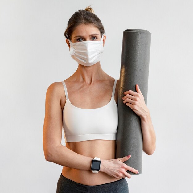 Vista frontal de la mujer con máscara médica sosteniendo estera de yoga