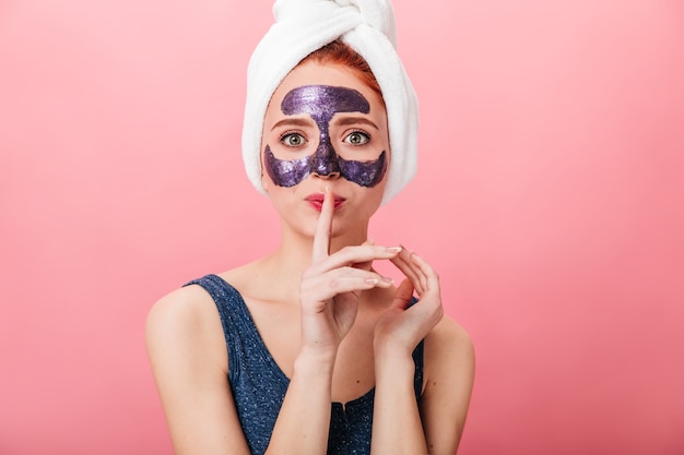 Vista frontal de la mujer con mascarilla mostrando signo de silencio. Disparo de estudio de señorita en toalla posando sobre fondo rosa durante el tratamiento de cuidado de la piel.