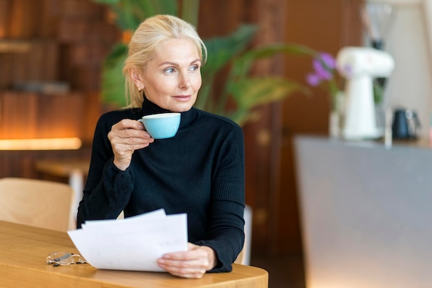 Vista frontal de la mujer mayor en el trabajo tomando café y leyendo periódicos