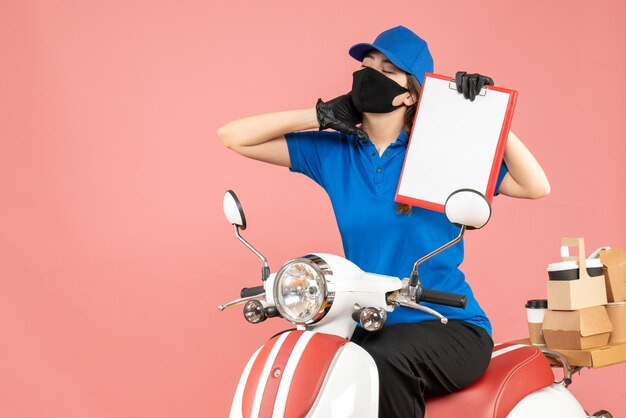 Vista frontal de la mujer de mensajería cansada con máscara médica y guantes sentado en scooter sosteniendo hojas de papel vacías entregando pedidos sobre fondo melocotón pastel