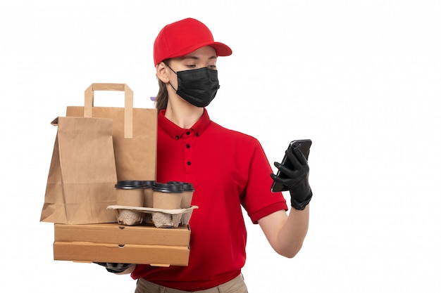 Una vista frontal mujer mensajero en carpa roja camisa roja guantes negros y máscara negra con paquetes de alimentos y tazas de café usando un teléfono en el servicio blanco