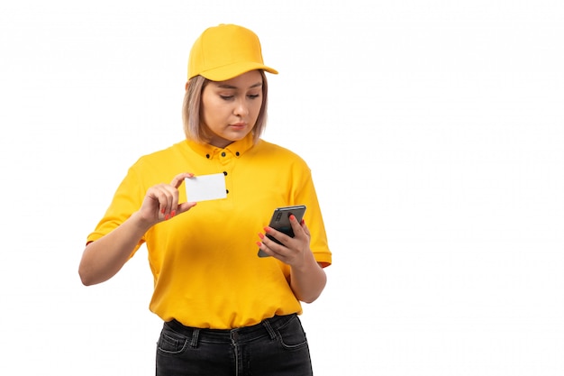 Una vista frontal mujer mensajero joven en camisa amarilla gorra amarilla y jeans negros con tarjeta blanca y teléfono inteligente en uniforme blanco