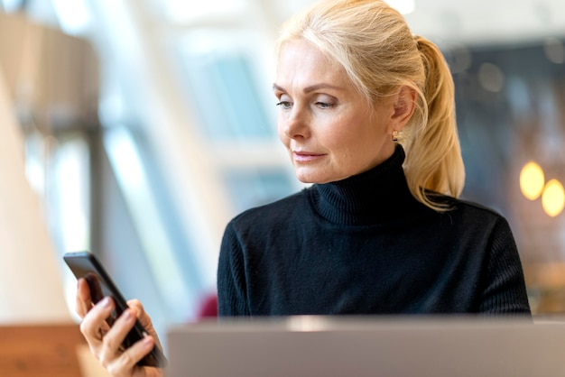 Vista frontal de la mujer de negocios mayor que trabaja en la computadora portátil y mirando el teléfono inteligente