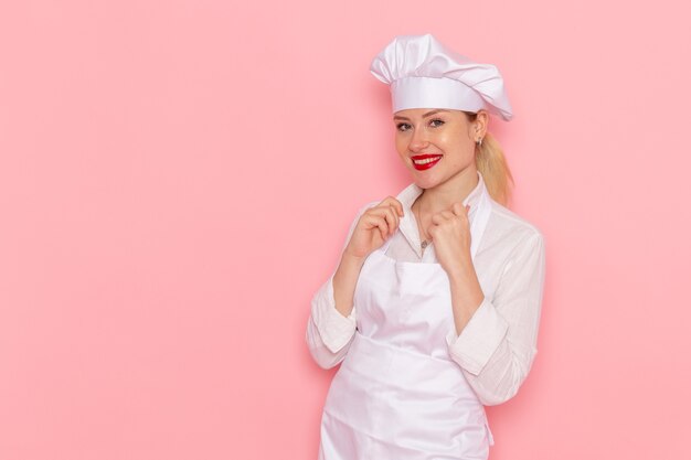 Vista frontal mujer pastelera en ropa blanca sonriendo y posando en la pared de color rosa claro cocinar confitería trabajo de pastelería dulce