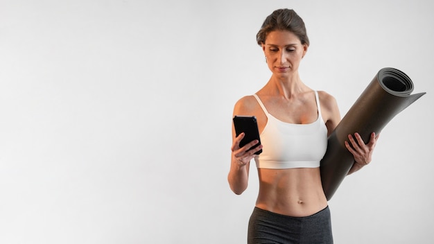 Foto gratuita vista frontal de la mujer que usa el teléfono inteligente mientras sostiene la estera de yoga con espacio de copia