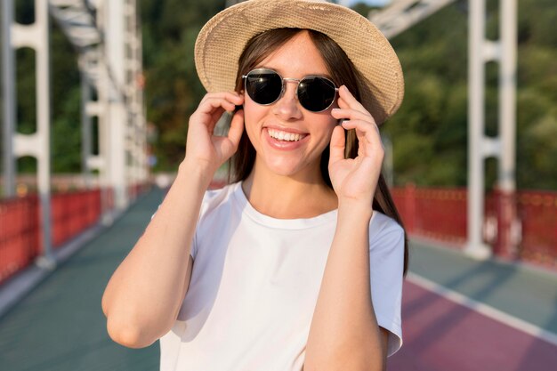 Vista frontal de la mujer que viaja sonriente en el puente con sombrero y gafas de sol