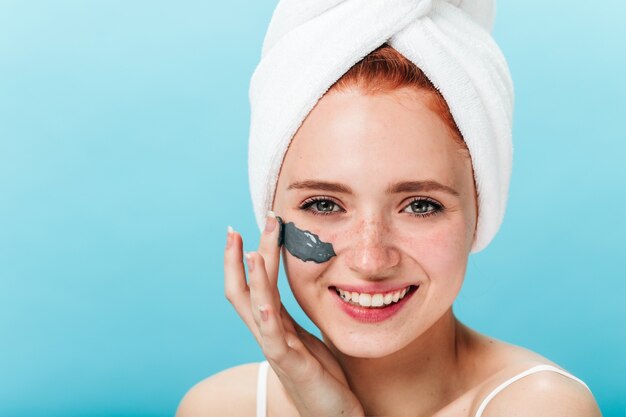 Vista frontal de la mujer riendo aplicando mascarilla. Foto de estudio de chica europea haciendo cuidado de la piel con una sonrisa.