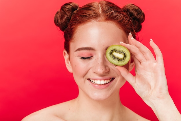 Vista frontal de la mujer riendo con kiwi. Foto de estudio de chica jengibre emocionada con frutas exóticas.