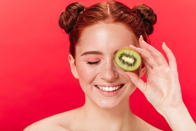 Vista frontal de la mujer riendo con kiwi. Foto de estudio de chica jengibre emocionada con frutas exóticas.