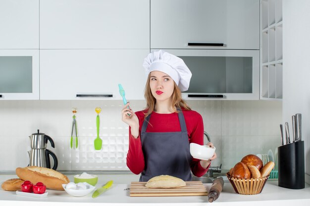 Vista frontal de la mujer rubia con sombrero de cocinero y delantal con tazón y cepillo en la cocina