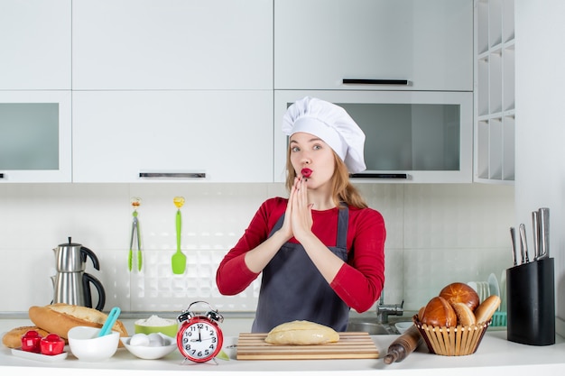 Vista frontal de la mujer rubia con sombrero de cocinero y delantal uniendo las manos en la cocina