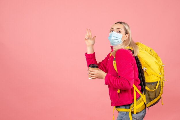 Vista frontal mujer rubia viajera con mochila amarilla con máscara sosteniendo la taza de café