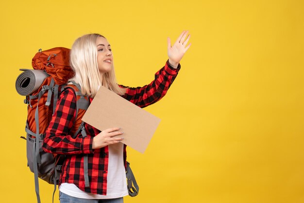 Vista frontal mujer rubia viajera con su mochila sosteniendo cartón saludando a alguien en la pared amarilla