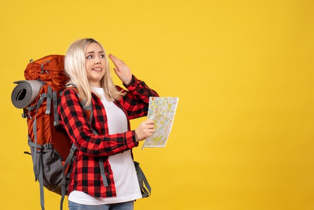Vista frontal mujer rubia viajera con su mochila sosteniendo el mapa de pie en la pared amarilla