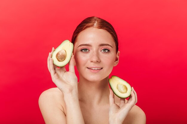 Vista frontal de una mujer sensual sosteniendo aguacate Foto de estudio de una chica jengibre sonriente aislada en un fondo rojo