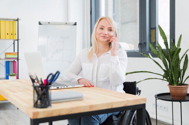 Vista frontal de la mujer en silla de ruedas posando mientras habla por teléfono en el trabajo