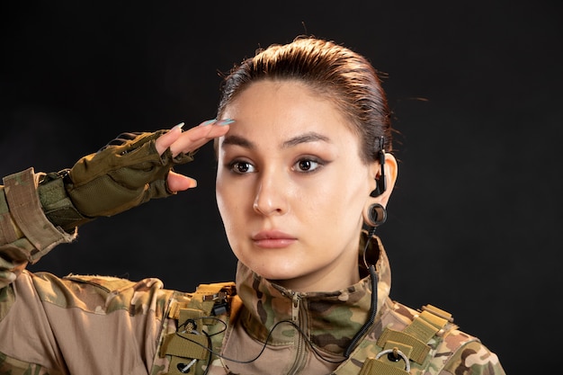 Vista frontal de la mujer soldado en camuflaje saludando en la pared negra