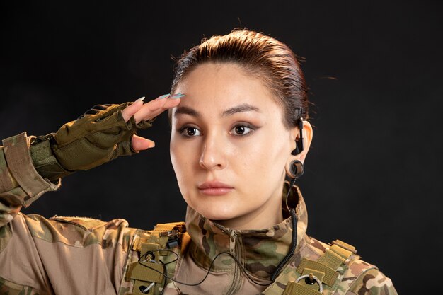 Vista frontal de la mujer soldado en camuflaje saludando en la pared negra