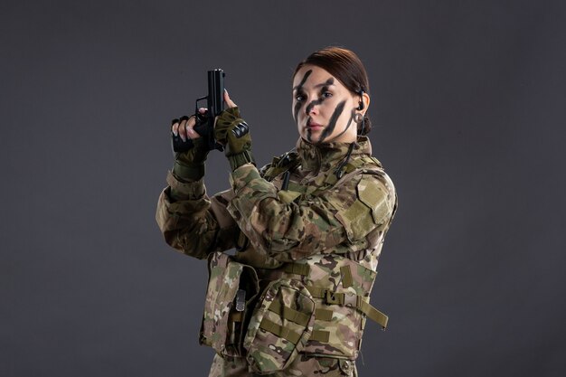 Vista frontal de la mujer soldado con pistola en pared oscura de camuflaje