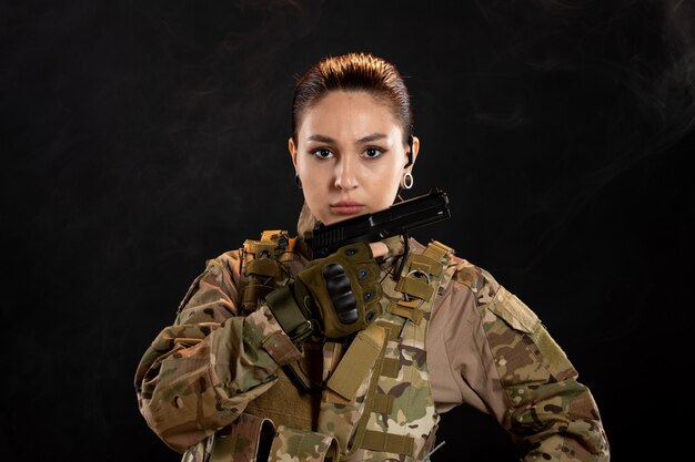 Vista frontal de la mujer soldado con pistola en uniforme sobre pared negra