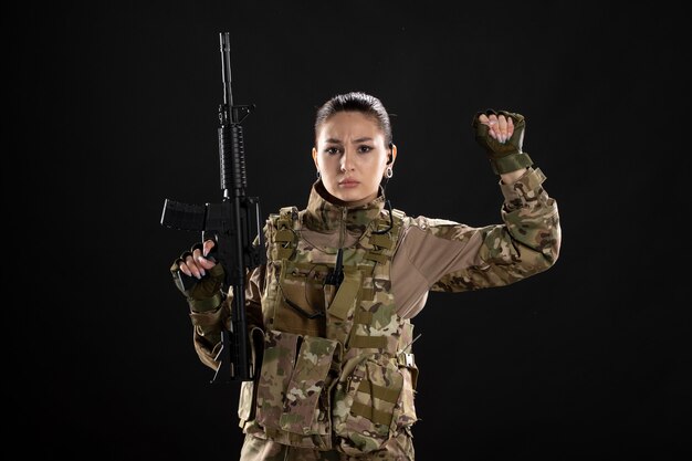 Vista frontal mujer soldado en uniforme con rifle en pared negra