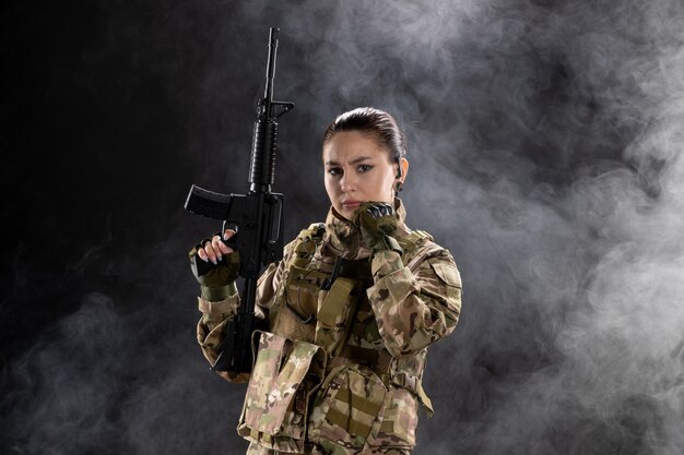 Vista frontal mujer soldado en uniforme con rifle en pared negra