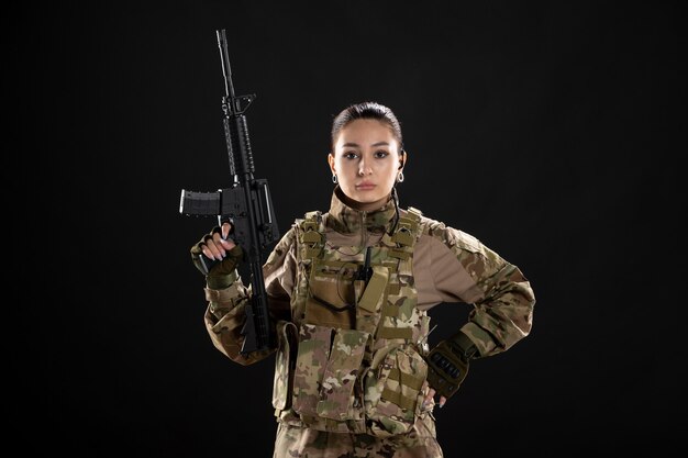 Vista frontal mujer soldado en uniforme con rifle en pared negra