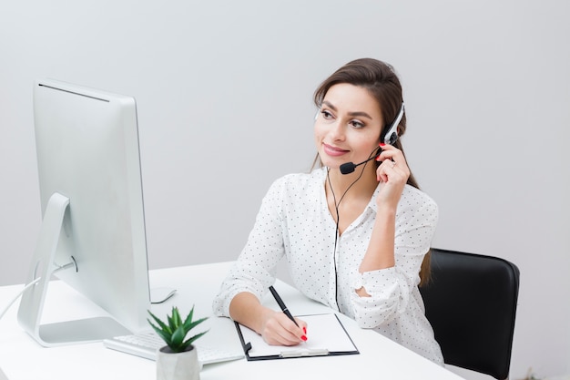 Vista frontal de la mujer sonriente escribiendo algo mientras habla por auriculares