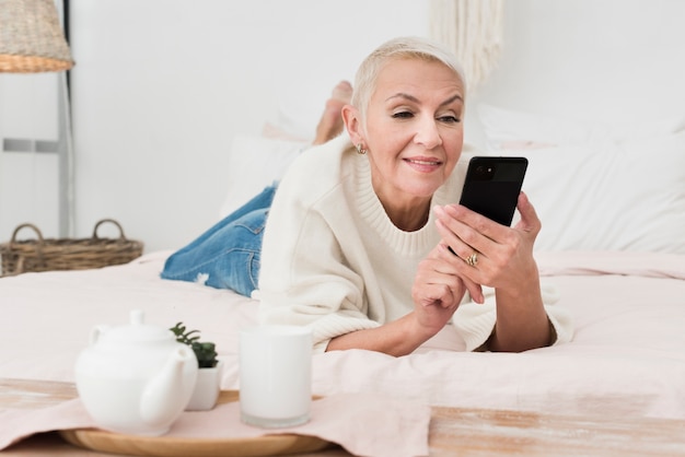 Vista frontal de la mujer sonriente madura en la cama con smartphone