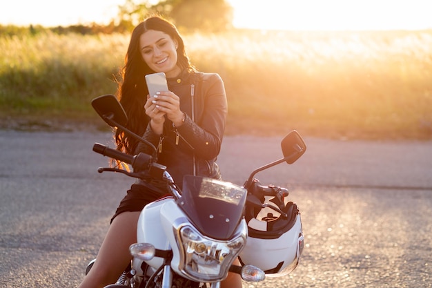 Vista frontal de la mujer sonriente mirando el teléfono inteligente mientras está sentado en su motocicleta