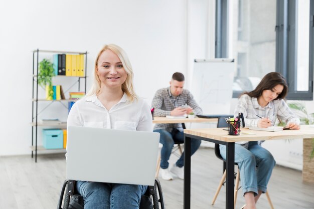 Vista frontal de la mujer sonriente en silla de ruedas posando con laptop en el trabajo