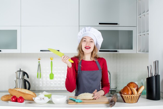 Vista frontal de la mujer sonriente con sombrero de cocinero y delantal sosteniendo un cuchillo en la cocina