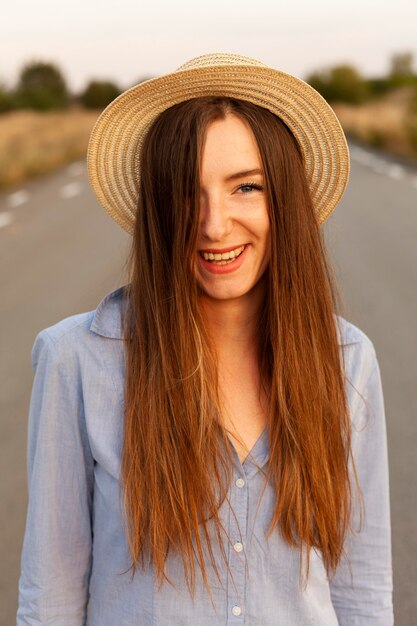 Vista frontal de la mujer sonriente con sombrero posando al atardecer en la carretera