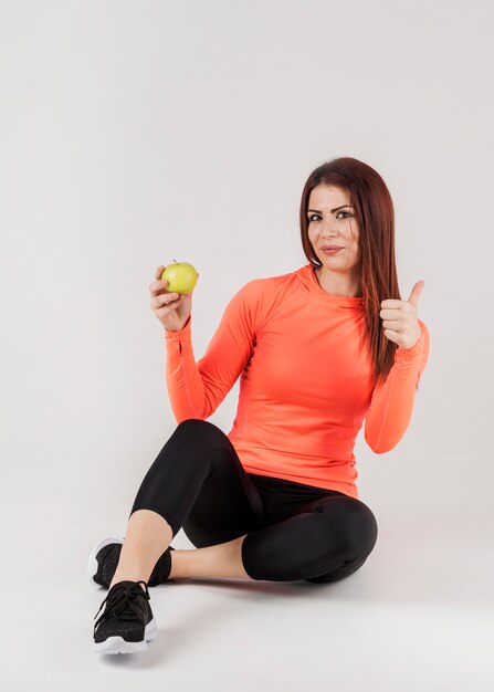 Vista frontal de la mujer en traje de gimnasio dando pulgares mientras sostiene la manzana