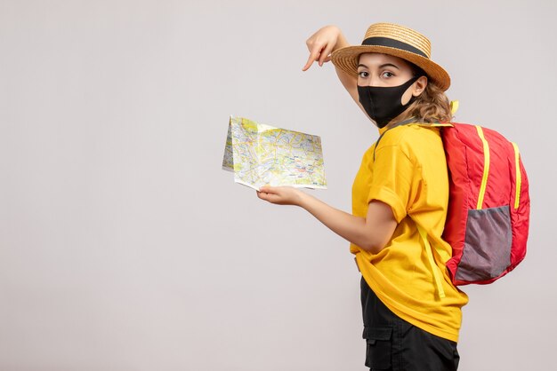 Vista frontal de la mujer viajera con máscara negra apuntando al mapa en la pared blanca