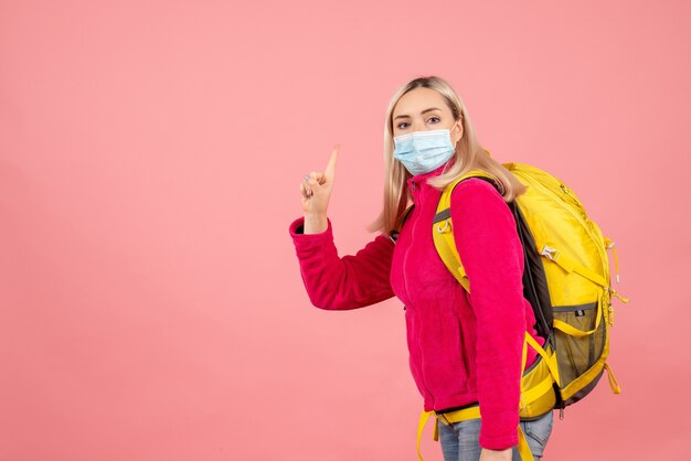 Vista frontal mujer viajero con mochila amarilla con máscara apuntando con el dedo hacia arriba