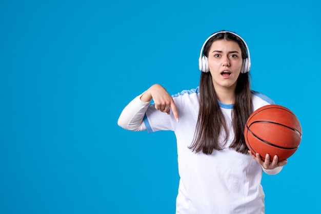 Vista frontal de las mujeres jóvenes con auriculares sosteniendo baloncesto en la pared azul