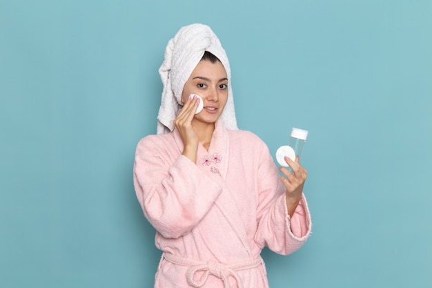 Vista frontal de las mujeres jóvenes en bata de baño rosa limpiando su rostro del maquillaje en el escritorio azul, limpieza, belleza, autocuidado, crema, ducha