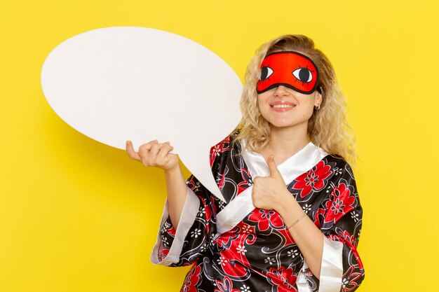 Vista frontal de las mujeres jóvenes en bata de noche sosteniendo un enorme cartel blanco sonriendo en la pared de color amarillo claro, modelo de cama de noche para dormir