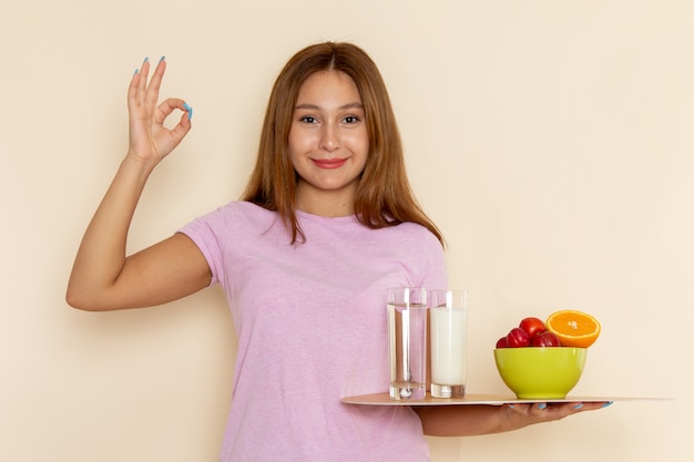 Vista frontal de las mujeres jóvenes en camiseta rosa y jeans con bandeja de frutas, leche y agua sonriendo en gris