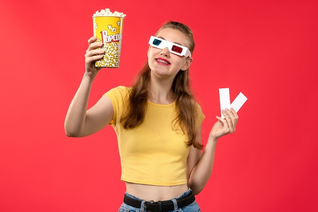 Vista frontal de las mujeres jóvenes en el cine con palomitas de maíz y entradas en gafas de sol d en la pared roja cine colores femeninos