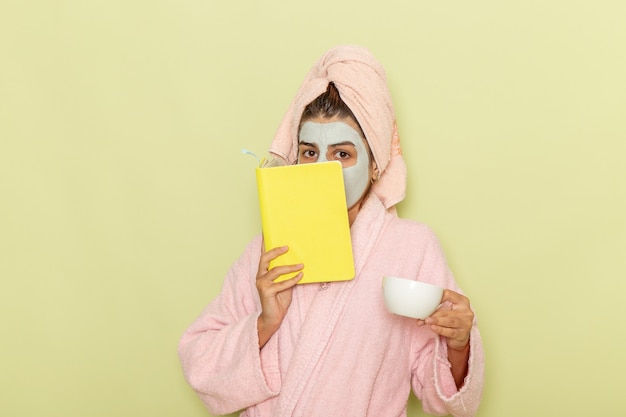 Vista frontal de las mujeres jóvenes después de la ducha en bata de baño rosa tomando café y leyendo el cuaderno en el escritorio verde