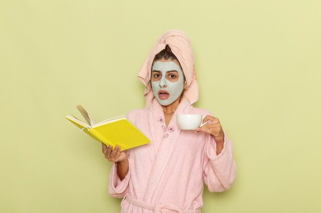 Vista frontal de las mujeres jóvenes después de la ducha en bata de baño rosa tomando café y leyendo el cuaderno en el escritorio verde