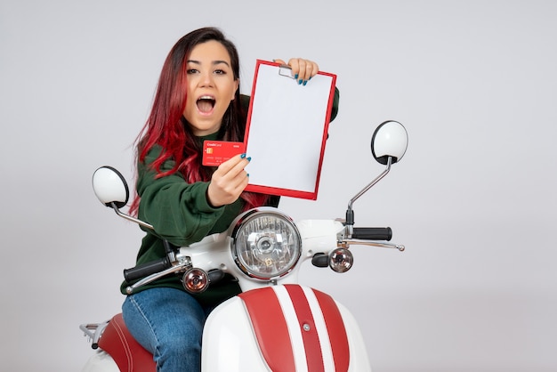 Vista frontal de las mujeres jóvenes en motocicleta sosteniendo una nota y una tarjeta bancaria en la pared blanca