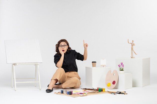 Vista frontal de las mujeres jóvenes sentados dentro de la habitación con caballete y pinturas en el piso blanco color arte dibujar cuadro pintor dibujo del artista